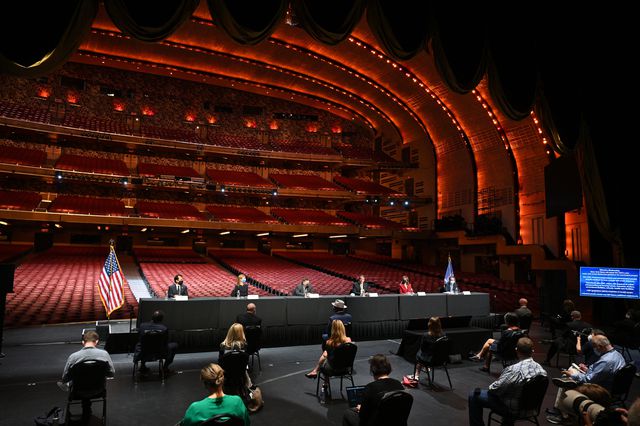 Governor Cuomo, James Dolan, Jane Rosenthal, and other members of Cuomo's staff with reporters on the stage of Radio City Music Hall, with the art deco flourishes of the venue seen in the background